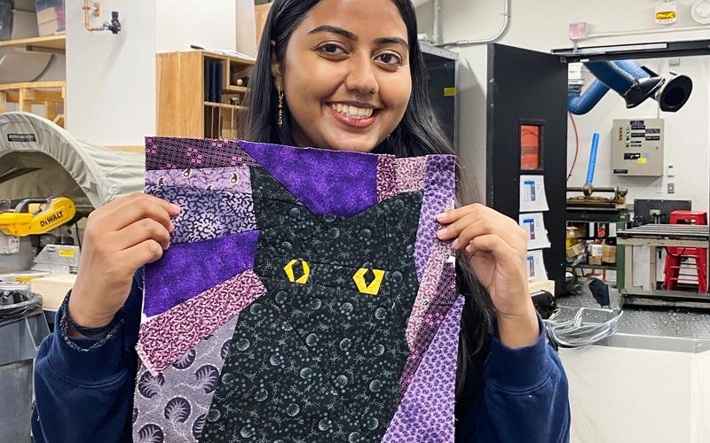 Person holding up a finished quilt block featuring a black cat with bright yellow eyes, surrounded by purple, lavender, and patterned fabric shapes. The background appears to be a makerspace workshop.